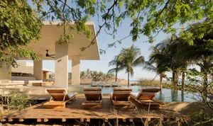 A modern residential outdoor area featuring an infinity pool, wooden deck chairs, and concrete structures surrounded by palm 