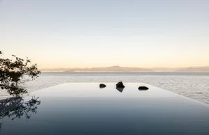 A minimalist infinity pool with water extending to the horizon, reflecting the sky and distant mountains, surrounded by natur