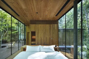 A modern minimalist bedroom with a white bed, wooden flooring, and a large glass wall overlooking trees.