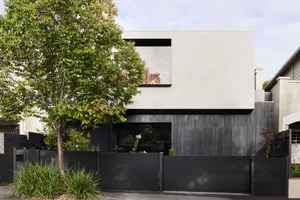 A modern residential house with a concrete exterior, surrounded by greenery and a black fence.