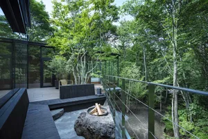 A modern outdoor space featuring a wooden deck, glass railings, and a concrete fire pit surrounded by lush greenery.