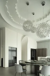 Modern dining room featuring a metal chandelier with geometric patterns, grey upholstered chairs, and a round granite table.