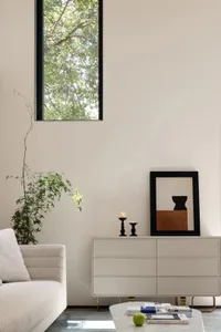 Minimalist living room featuring neutral-colored furniture and decor, with a tall window showcasing greenery outside.