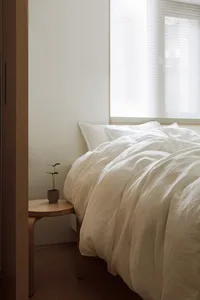 A minimalist bedroom with a bed covered in beige linen and a small wooden side table.