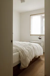 A minimalist bedroom with white walls and a wood floor, featuring a bed with a white comforter and a small bedside table with