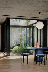 A modern, minimalist dining room with a large round table, stools, and a concrete and glass structure visible through large w