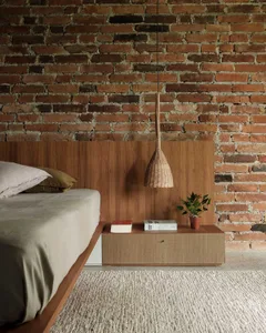 A minimalist bedroom with a wooden bed frame, brick wall, woven pendant light, and a nightstand with books and a plant.
