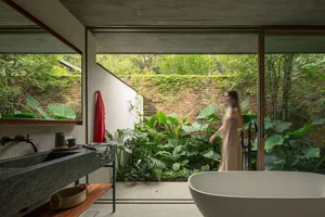 Modern bathroom with concrete walls, large windows, and green tropical plants outside, featuring a freestanding white bathtub