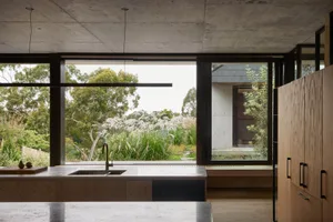 A modern kitchen with concrete and wooden elements features a sink and a view of a garden through large windows.
