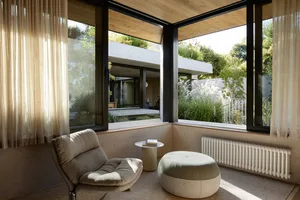 A modern minimalist living space with a comfortable armchair, round ottoman, and large windows allowing natural light in, ove