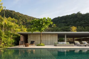 Modern residential poolside area with concrete structure, wooden furniture, and lush greenery.
