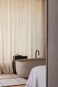 Minimalist bathroom featuring a marble bathtub, linen curtains, and porcelain fixtures.