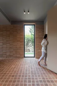 A person in a minimalist, brick-clad residential interior looking out through a tall, narrow window.