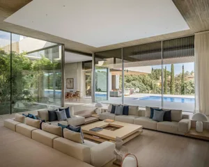 Modern living room featuring glass walls and wooden furniture, with a large open space and a view of a pool and greenery outs
