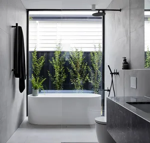 Modern minimalist bathroom with concrete walls, white bathtub, and green plants in front of a black wooden fence.