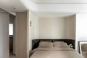 A minimalist bedroom featuring a bed with a black upholstered headboard and beige bedding, alongside a wooden headboard panel