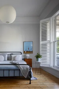 Modern minimalist bedroom featuring a metal-framed bed on a wooden floor with a white plaster ceiling and white window shutte