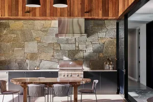 A modern kitchen with stone and concrete walls, a teak wood table, and a stainless steel grill and hood.