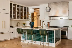 A minimalist kitchen with white marble countertops, brass hardware, and green tile accents, featuring white cabinets, pendant