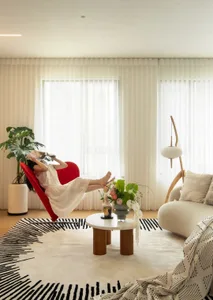 A modern minimalist living room with wooden furniture, white curtains, and a red accent chair.