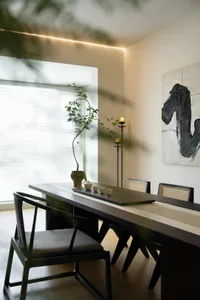 A minimalist dining room with a long wooden table and chairs, placed near a large window with a plant and candles.
