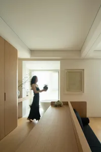 A minimalist residential interior featuring white walls and white wood flooring, with a person holding a vase.