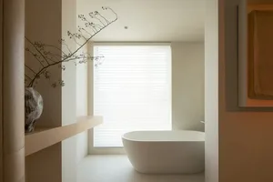 Modern bathroom featuring a freestanding white bathtub, a window with closed blinds, and a vase with dried plants on a shelf.