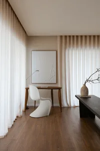 Modern minimalist office space featuring a desk, chair, and wood table, with curtains and a painting on the wall.
