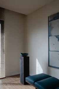 A modern living room featuring hardwood floors, a blue upholstered bench, and a tall black pedestal with a decorative bowl on