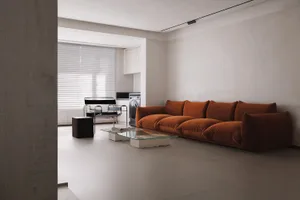 A minimalist living room with a brown velvet couch, a glass coffee table, and a kitchen area in the background.