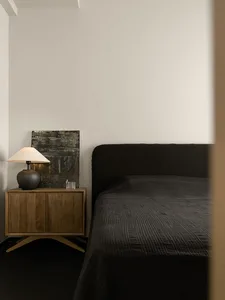 Modern, minimalist bedroom featuring dark textured bedding on a bed with a simple, upholstered headboard, accompanied by a wo