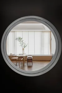 A minimalist living room with a large window featuring wood frames and blinds, a wooden table and chairs, and a wooden floor 