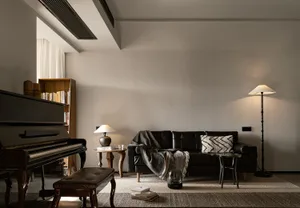 A modern living room featuring a black leather sofa, a floor lamp, and a piano with books and a lamp on a side table.