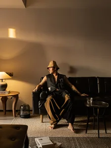 A man wearing a tan bucket hat and a black shirt with a white patch is sitting on a black leather sofa in a living room with 