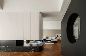 A modern minimalist living room featuring a white couch, blue and white rug, and a black marble coffee table. The space is il