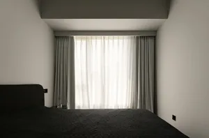 A minimalist bedroom with black furniture and grey walls, featuring white curtains and a window.