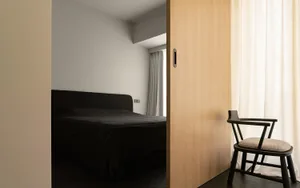 A modern minimalist bedroom with a dark bed, white curtains, and a wooden sliding door.