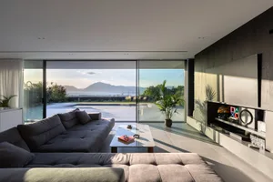 A modern minimalist living room with large glass walls and concrete flooring, featuring a spacious sofa and a sleek, minimali