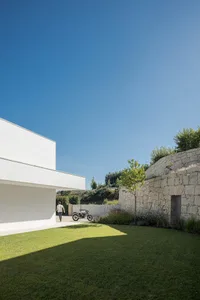 A minimalist home with white concrete walls and stone accents, featuring greenery and a clear blue sky.