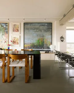 Modern dining room featuring a wooden table with glass top, a black pedestal table, and chairs with white cushions, accompani