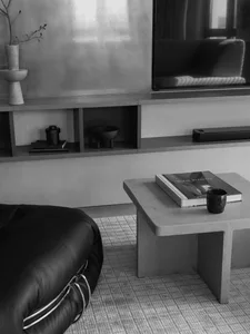 A modern minimalist living room with a concrete coffee table, a black leather ottoman, and a flat-screen TV mounted on the wa