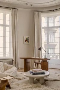 A modern living room featuring a white sofa, round table with books, wooden desk with lamp, and large windows with minimalist