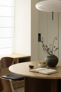 A cozy modern study space featuring wooden furniture, a vase with dried flowers, and an open book on a round wooden table.