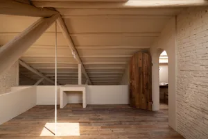 Minimalist interior space with exposed brick and plaster walls, wooden beams on the ceiling, and a wooden door.