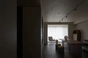 A modern living room featuring a wooden floor, minimalist furniture, and large windows with white curtains.
