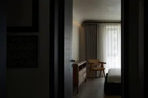 A modern, minimalist bedroom with a wooden chair and a large window with curtains.