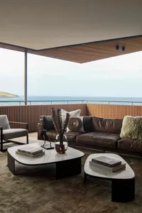 Modern industrial-style living room with a brown leather sofa, wooden coffee table, gray armchair, and large ocean view.