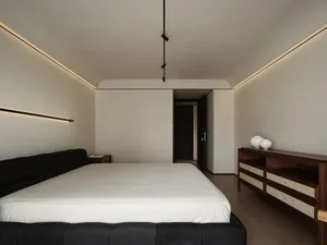 A modern minimalist bedroom features a bed with a dark headboard, white bedding, and neutral-toned walls and flooring.