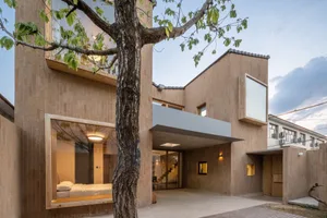 A modern residential building constructed with wood materials, featuring a large window and a tree in the foreground.