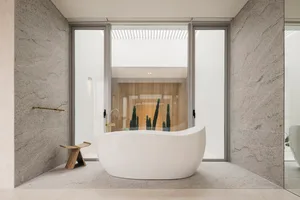A minimalist bathroom with a white bathtub, wooden stool, and large windows showing a cactus plant.